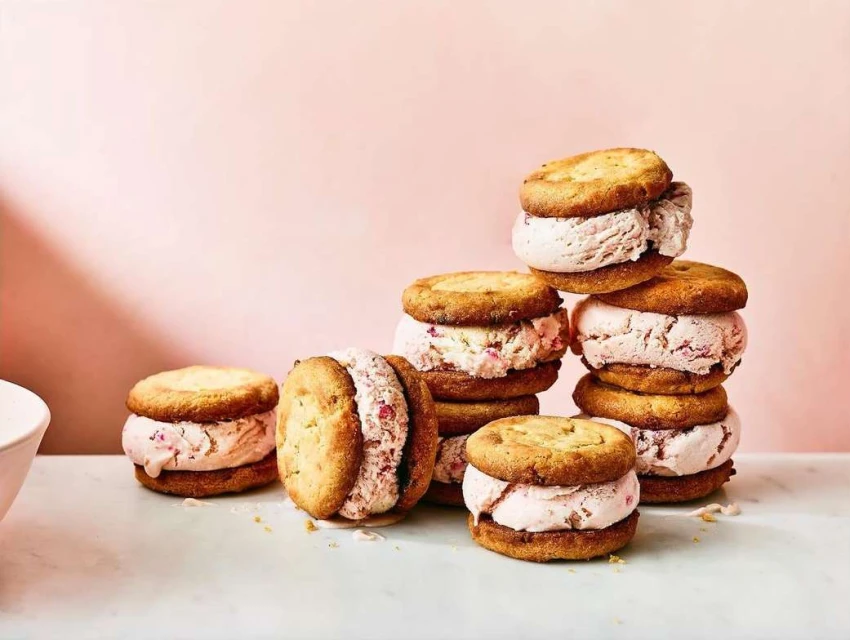Stack of ice cream sandwiches with round cookies and light pink ice cream filling against a pink background; food and beverage.