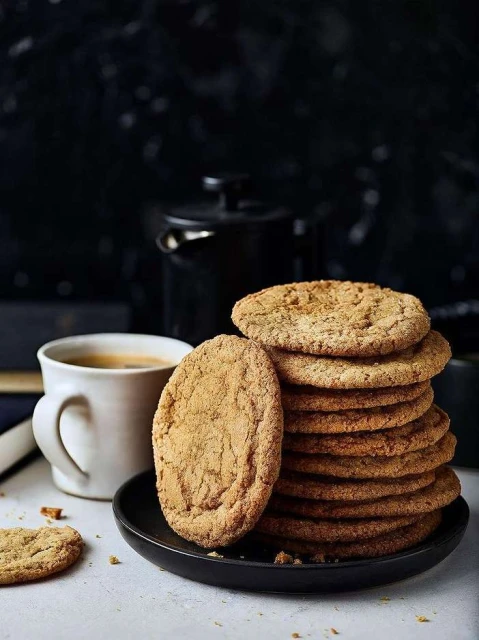 Stack of cookies on a black plate, next to a white cup of coffee and a black coffee pot, against a dark background.