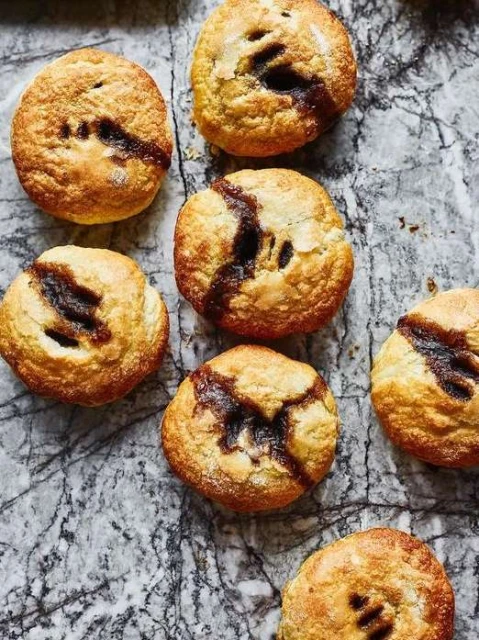 Overhead shot of seven golden-brown pastries with dark filling on a grey marble surface, showcasing baked goods and food photography.