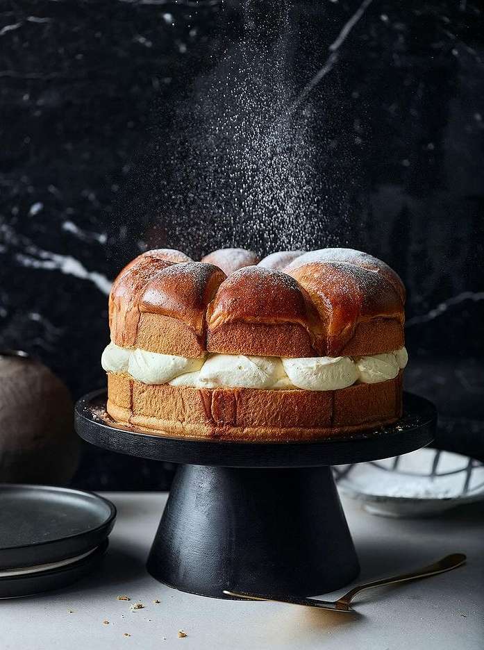 Brioche cake covered in powdered sugar and filled with cream, sitting on a black cake stand, against a dark marble background.