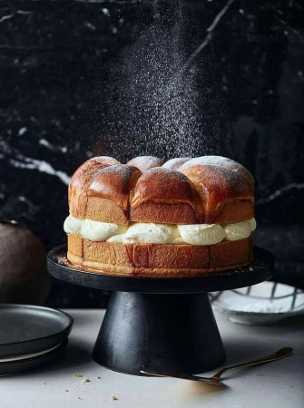 Brioche cake covered in powdered sugar and filled with cream, sitting on a black cake stand, against a dark marble background.