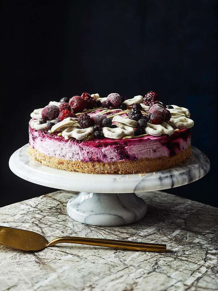 Overhead shot of a mixed berry cheesecake topped with meringue pieces and drizzled syrup on a marble stand, presented on a marble surface.