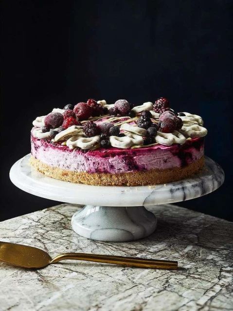 Overhead shot of a mixed berry cheesecake topped with meringue pieces and drizzled syrup on a marble stand, presented on a marble surface.