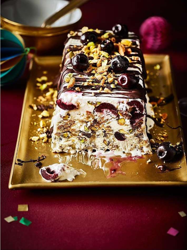 Eye-level shot of a semifreddo dessert topped with cherries and pistachios, presented on a gold tray against a red background.