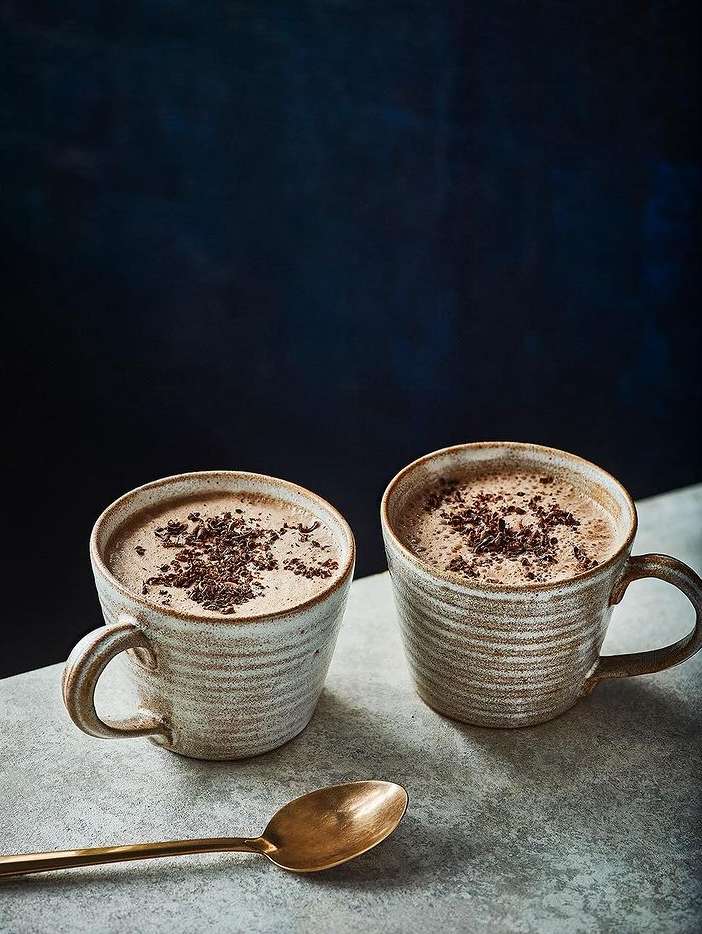 Two mugs of cocoa sit on a gray surface with a golden spoon, set against a dark blue backdrop with a diffused light source.