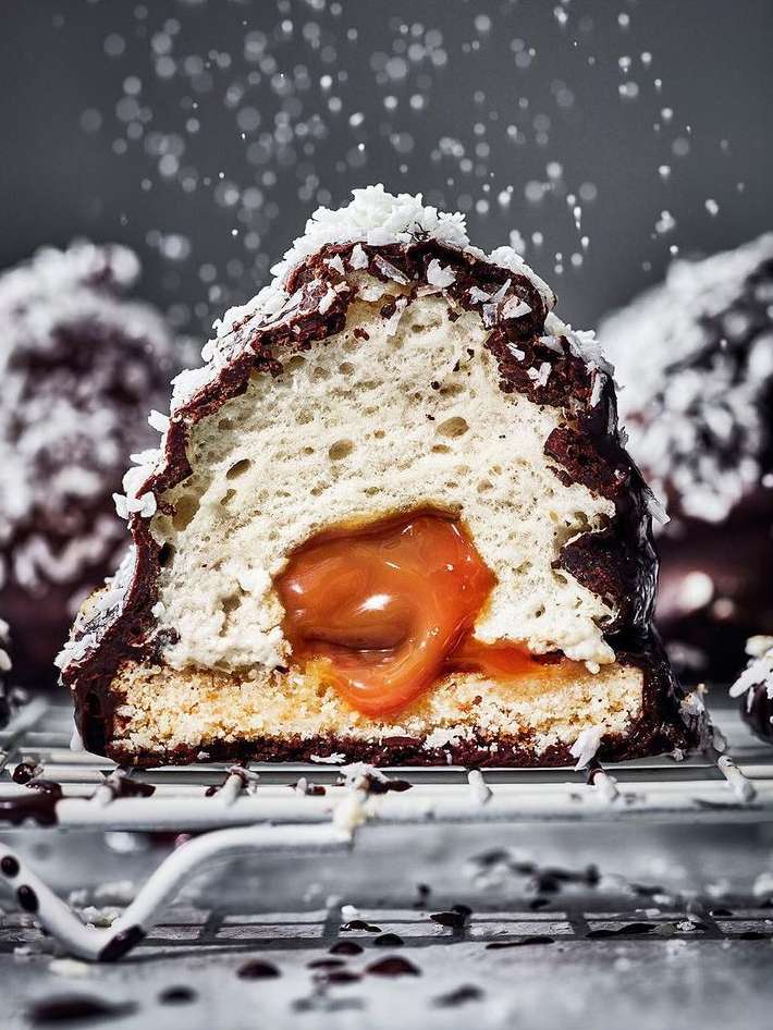 Close-up of a halved chocolate-covered marshmallow treat with caramel filling, topped with shredded coconut, on a wire rack.