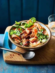 Bowl of noodle soup with chicken and vegetables on a wooden cutting board with a spoon alongside.