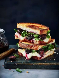 Two stacked toasted sandwiches filled with melted cheese, fresh blackberries, and green herbs on a dark cutting board.