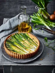 Leek tart on a dark ceramic plate with a glass oil bottle, fresh green onions, and eggs in London by Martin Poole