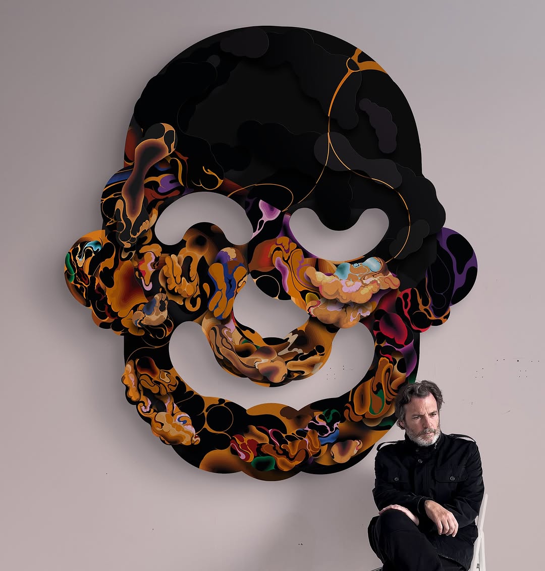 Man seated beside abstract artwork. Abstract face-like art with swirling colors on wall. Indoor shot with neutral lighting.