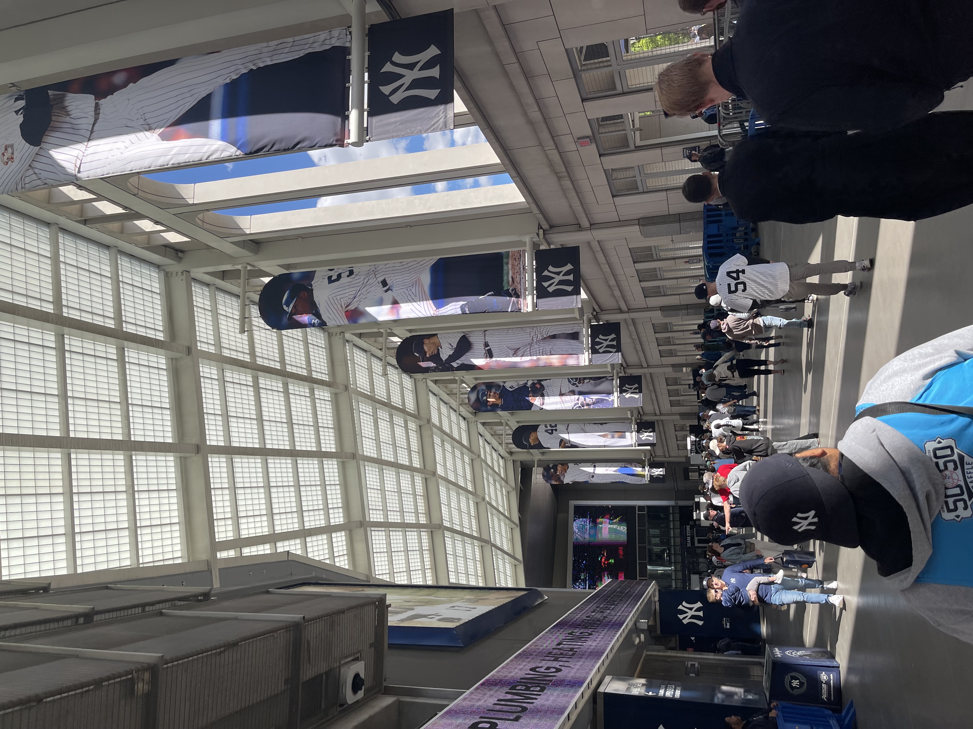 Crowd walking through a stadium concourse with large vertical baseball player banners hanging from a high glass roof.
