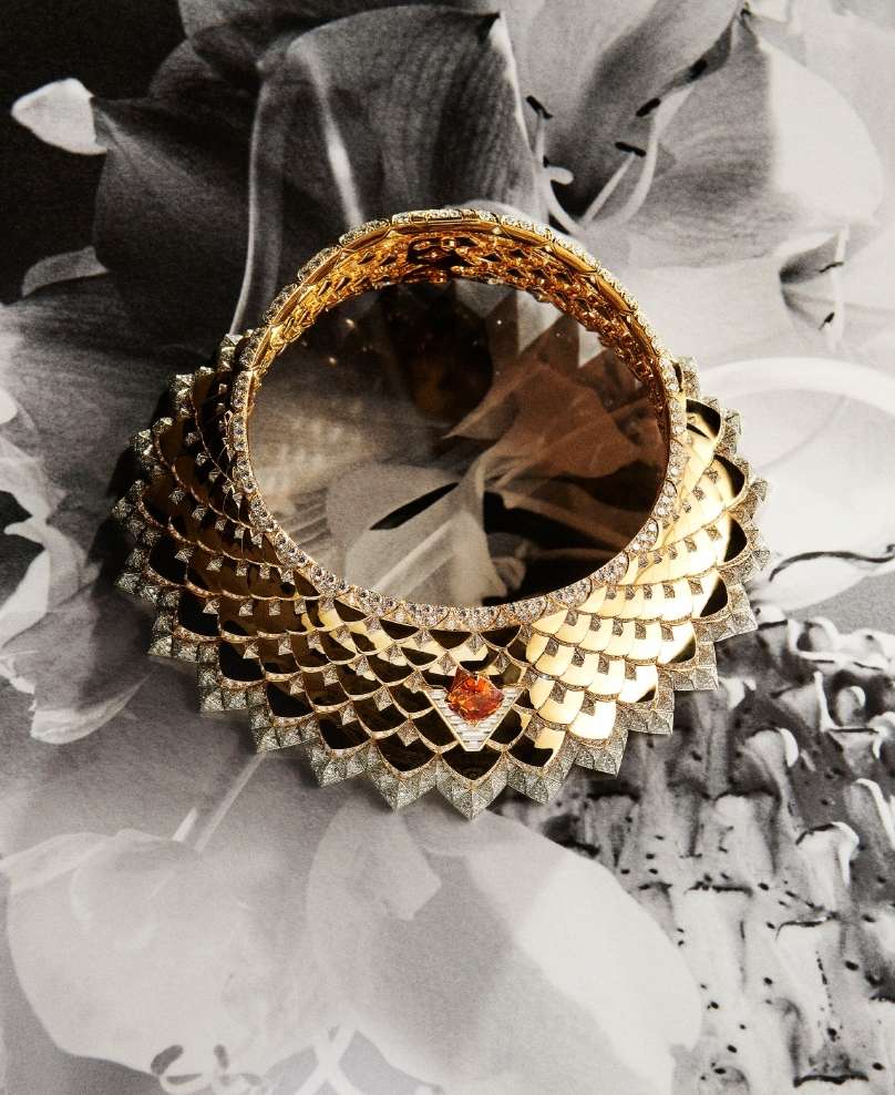 Elegant gold and diamond collar necklace featuring intricate scale design and orange gemstone, set against floral monochrome backdrop.