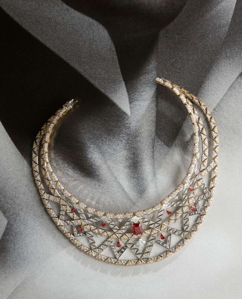 Close-up of a diamond-encrusted collar necklace with rubies, displayed against a blurred grayscale background of a draped garment.