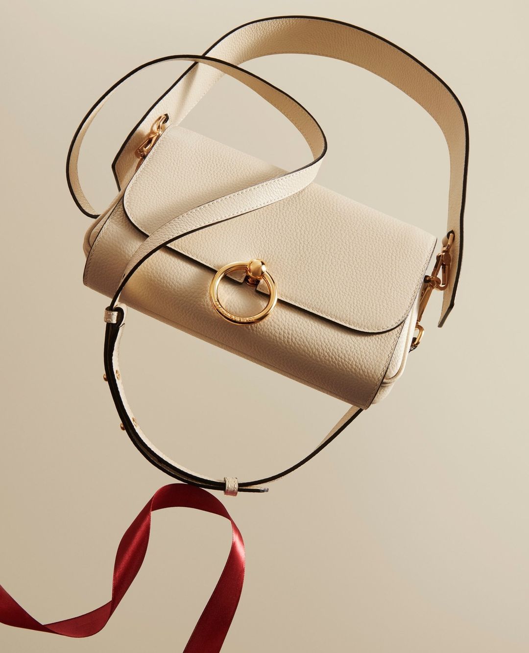 Cream-colored leather handbag with a wide strap and a gold ring detail, complemented by a burgundy ribbon, studio product shot.