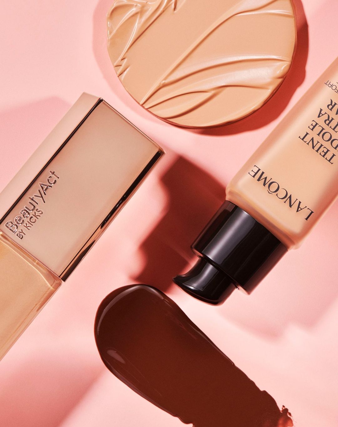 Close-up of Lancome foundation bottle, BeautyAct compact, and dark brown foundation swatch on light pink backdrop.