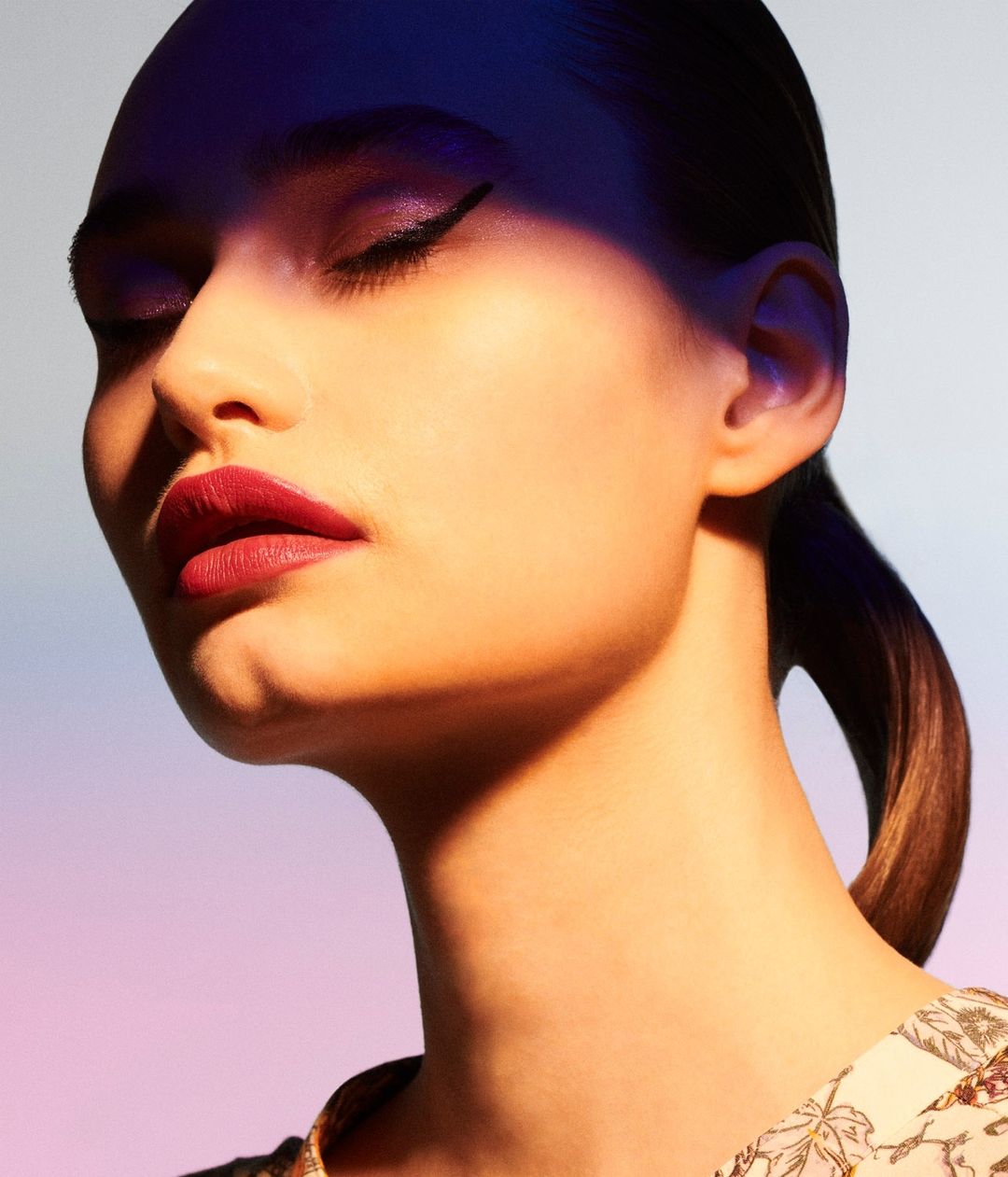 Close-up shot of a beautiful woman with eyes closed, wearing eye makeup and lipstick, against a gradient purple background in a beauty shot.