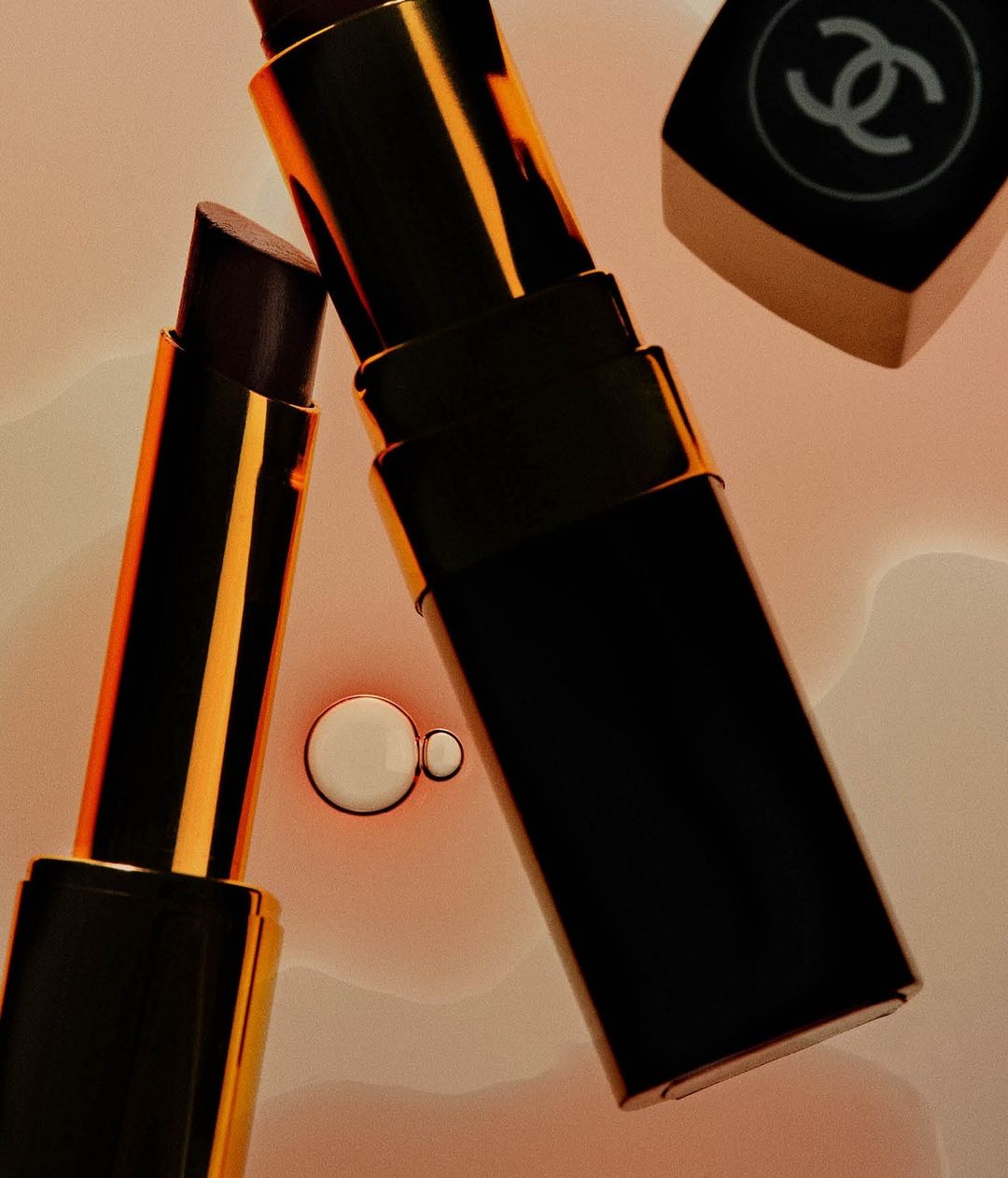 High-angle shot of black and gold Chanel lipsticks against a diffused peach background with floating droplets. Beauty and makeup.