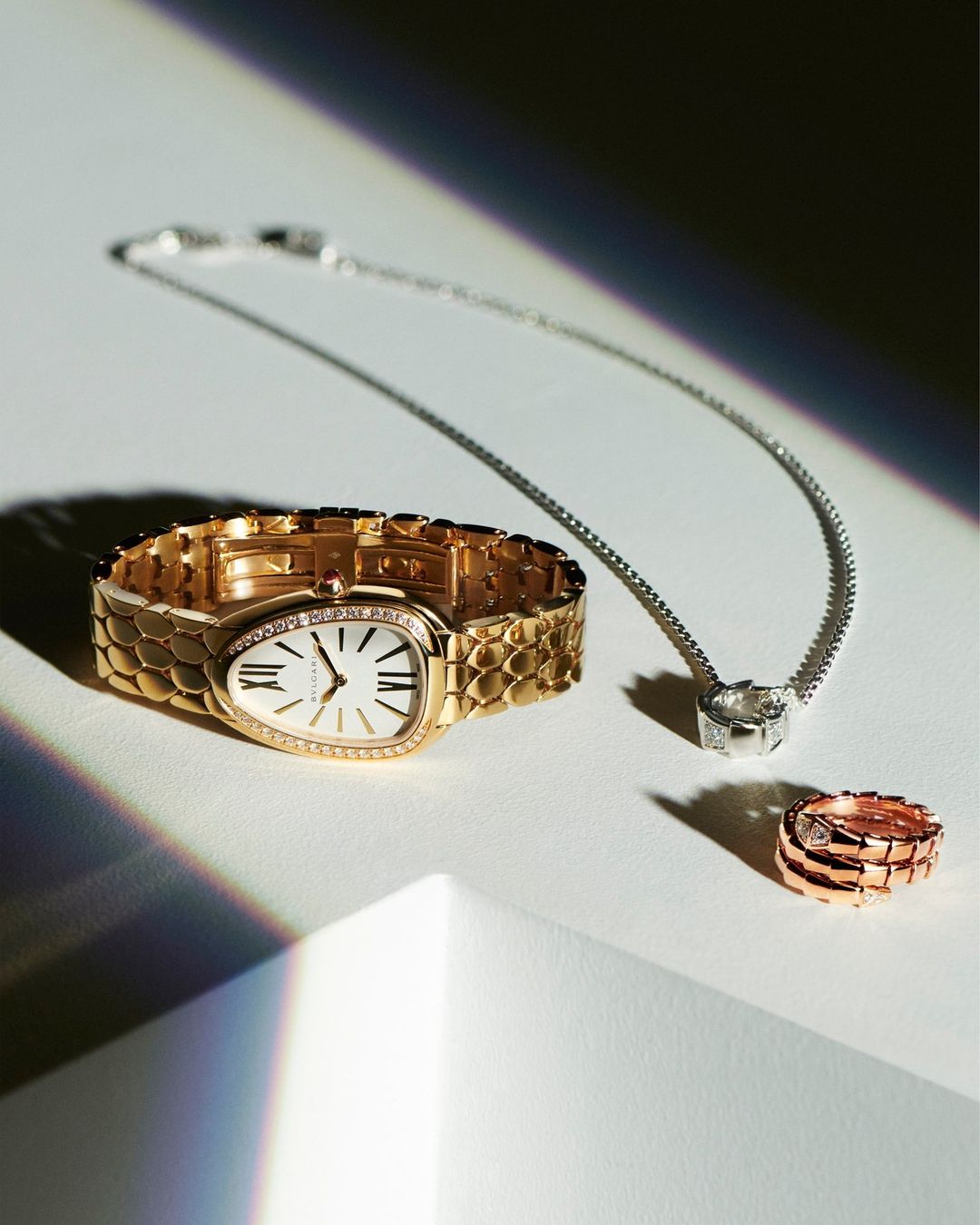 Still life shot of a gold watch, silver necklace, and rose gold ring on a white surface with a geometric shadow, highlighting the jewelry.