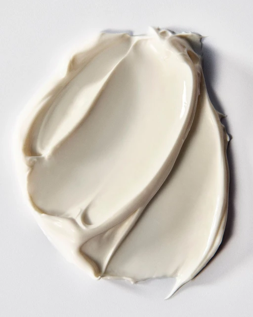 Close-up of a thick, light beige cream swatch with smooth, swirling textures on a white surface. Beauty product shot. 