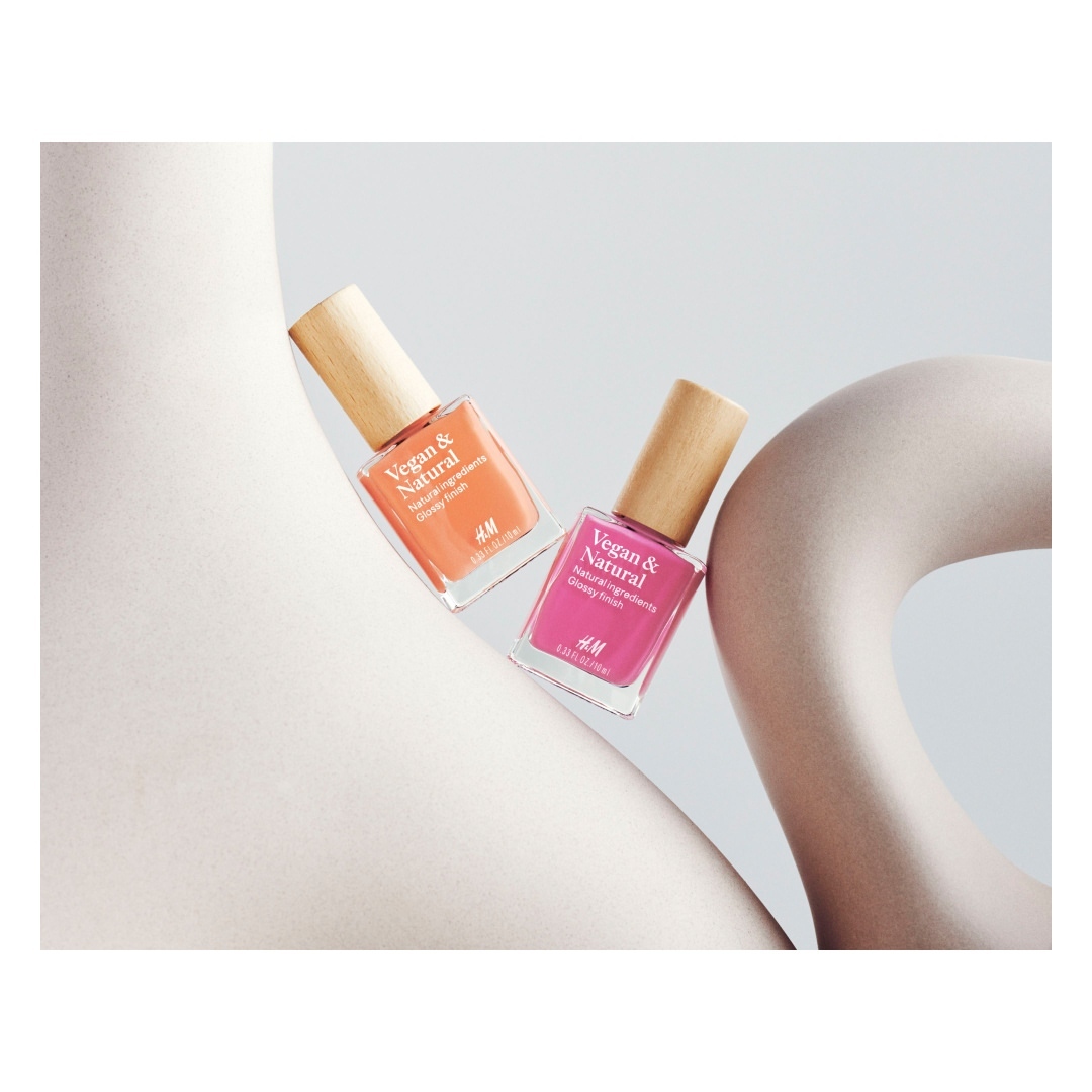 Two bottles of H&M Vegan & Natural nail polish, one peach and one pink, are displayed against a white abstract sculpture.