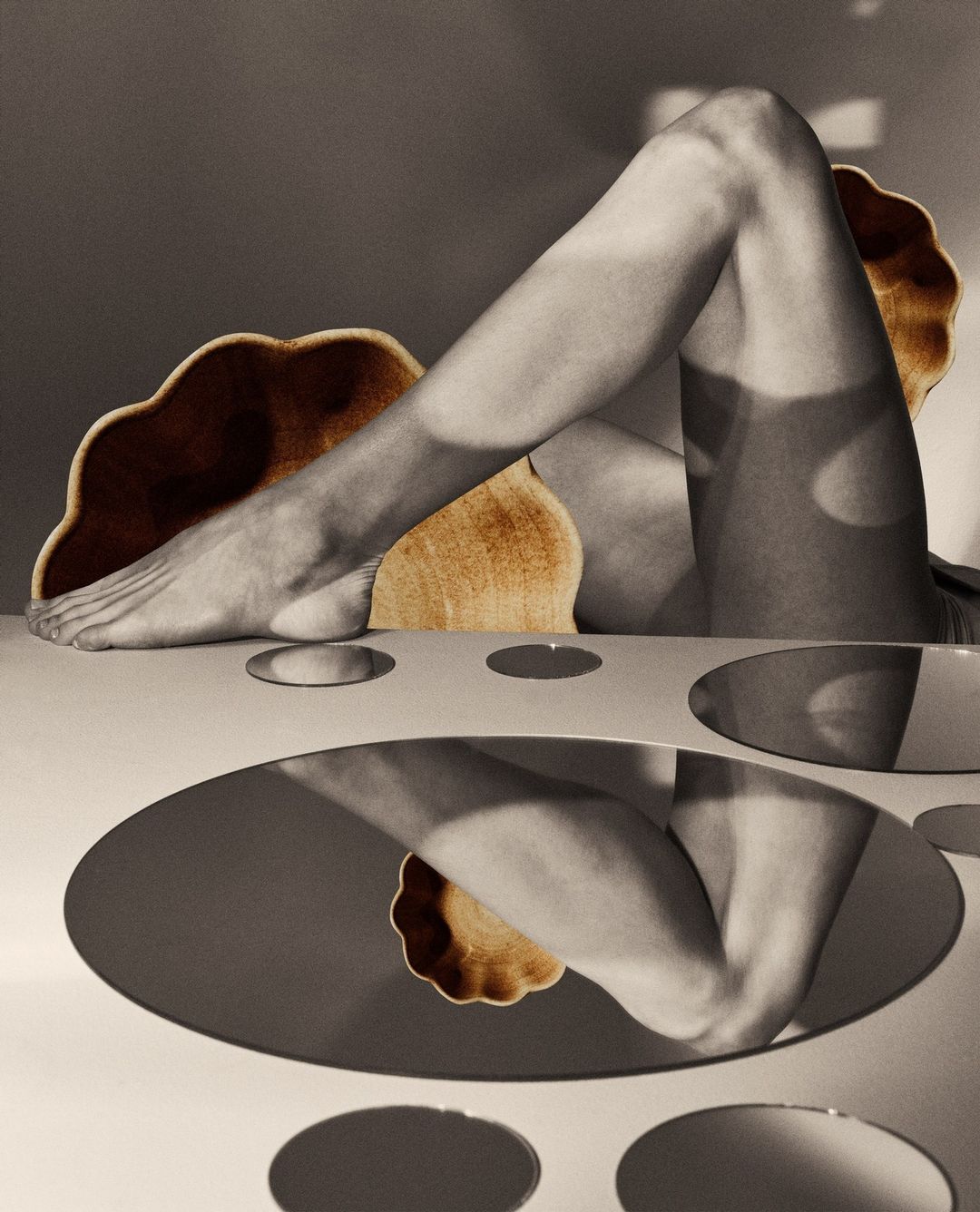 Monochromatic art photograph of a person's leg elevated on a table next to a stylized chair and reflective surfaces, creating a surreal studio scene.