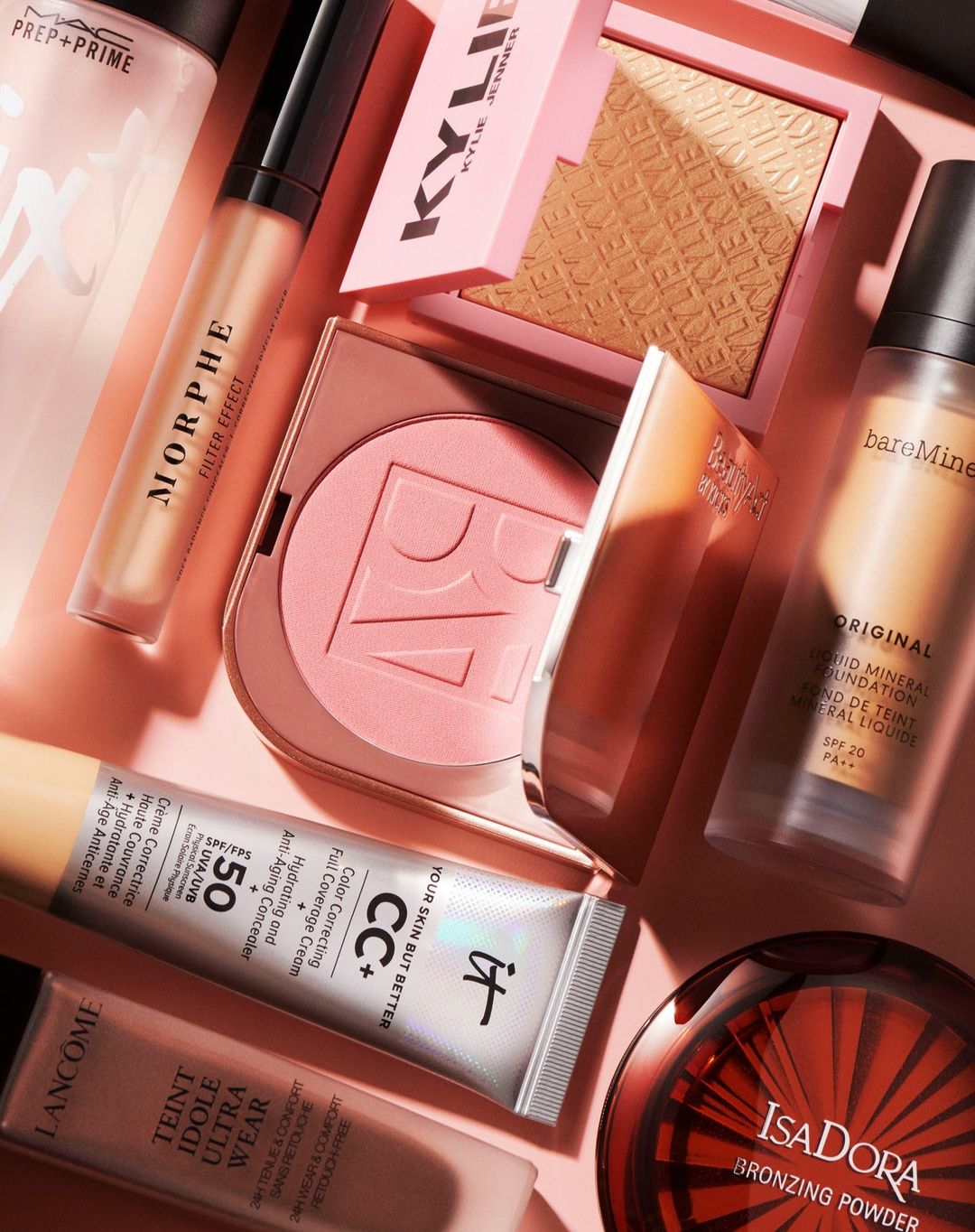 Still life of assorted makeup products including foundation, concealer, bronzer, and powder compacts in pink and neutral tones.