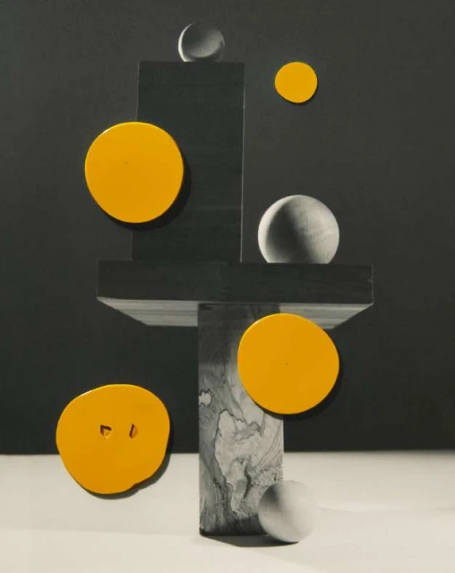 Abstract still life features yellow discs, gray spheres, and geometric gray and marble blocks against a dark gray backdrop and white base.