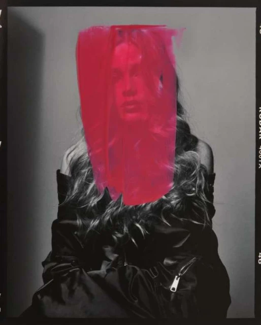 Fashion photo of an adult female with long blonde hair partially obscured by bright pink paint against a neutral gray background.