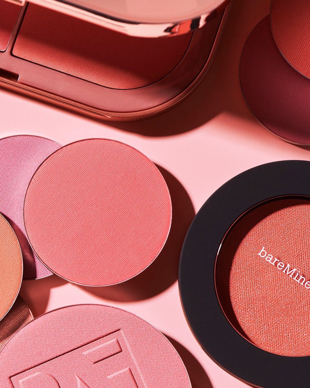Close-up of assorted cosmetic blush compacts and palettes in peach and pink tones against a soft pink background in a studio setting.