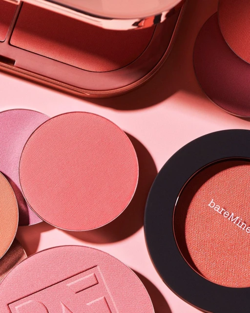 Close-up of assorted cosmetic blush compacts and palettes in peach and pink tones against a soft pink background in a studio setting.