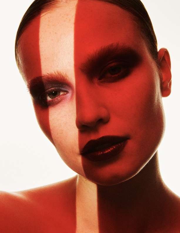 Close-up portrait of a female model with bold red makeup and a striking vertical stripe of light across her face, against a bright background.