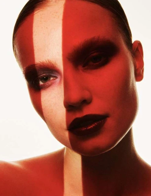 Close-up portrait of a female model with bold red makeup and a striking vertical stripe of light across her face, against a bright background.