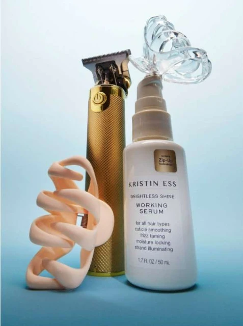Hair styling products on a light blue background feature a gold hair clipper and a white bottle of Kristin Ess Weightless Shine Working Serum.