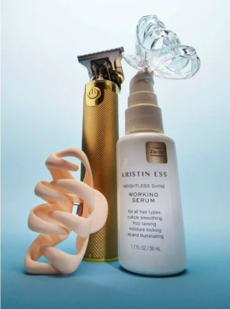 Hair styling products on a light blue background feature a gold hair clipper and a white bottle of Kristin Ess Weightless Shine Working Serum.