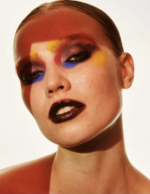 Close-up portrait of an adult woman with colorful makeup and glossy lips posing in front of a white background, highlighted with red light.