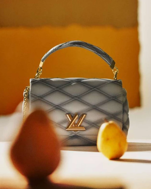Luxury gray quilted handbag with gold hardware and a subtle pear prop, set against a textured orange and cream backdrop.
