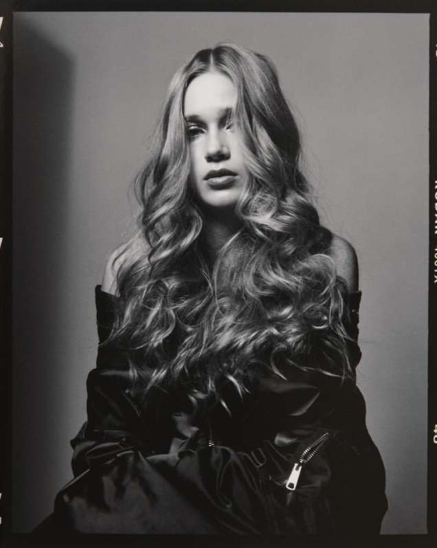 Black and white portrait of a young woman with long, wavy hair wearing a jacket and posing in front of a grey backdrop.