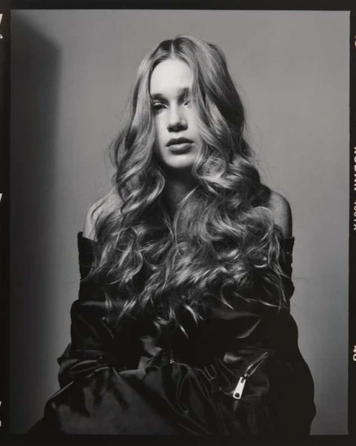 Black and white portrait of a young woman with long, wavy hair wearing a jacket and posing in front of a grey backdrop.