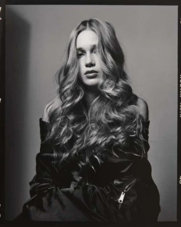 Black and white portrait of a young woman with long, wavy hair wearing a jacket and posing in front of a grey backdrop.