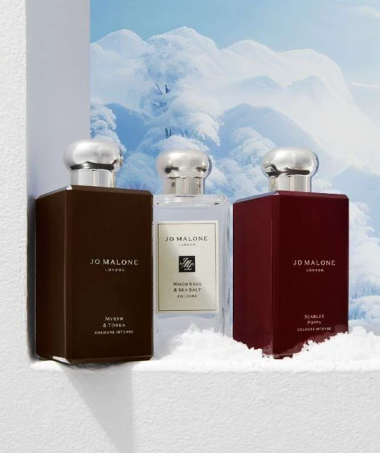 Three Jo Malone London cologne bottles in different colors displayed against a snowy mountain backdrop in a fragrance product shot.
