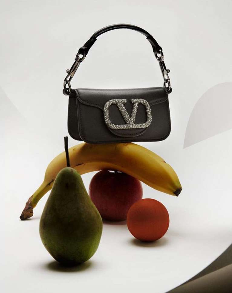 Still life featuring a black Valentino handbag with a jeweled logo, balanced on a yellow banana, green pear, and red apple against a white backdrop.