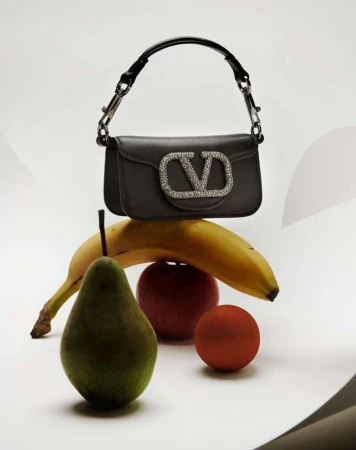 Still life featuring a black Valentino handbag with a jeweled logo, balanced on a yellow banana, green pear, and red apple against a white backdrop.