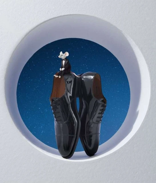 Conceptual fashion still life: shiny black patent leather men's dress shoes with a small grey mouse in a tuxedo perched on top.
