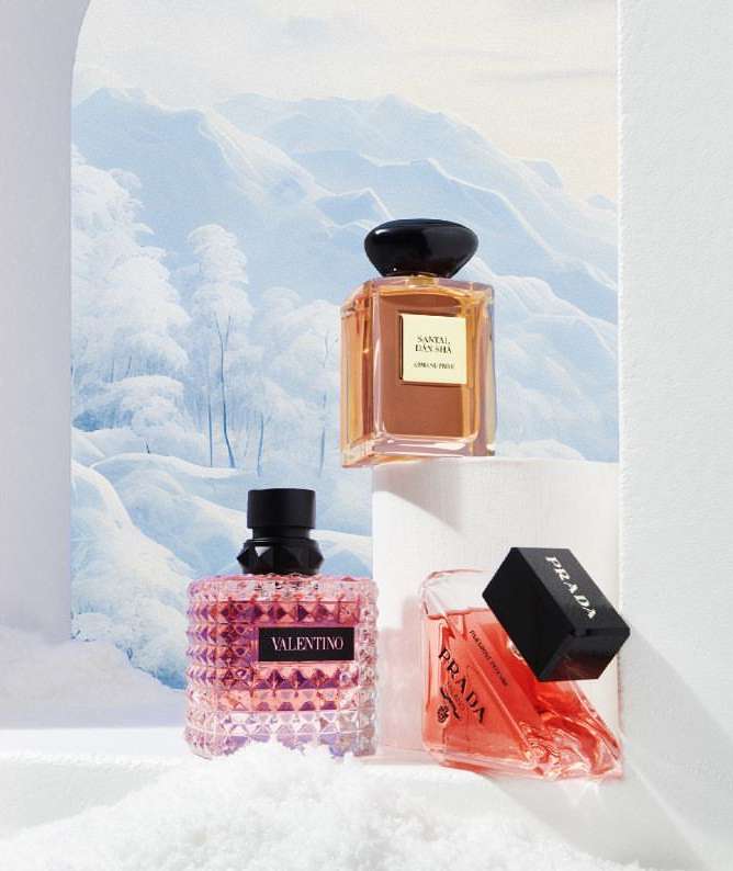 Three designer perfume bottles, Valentino, Armani, and Prada, sit on white surfaces against a soft blue and white mountain backdrop.