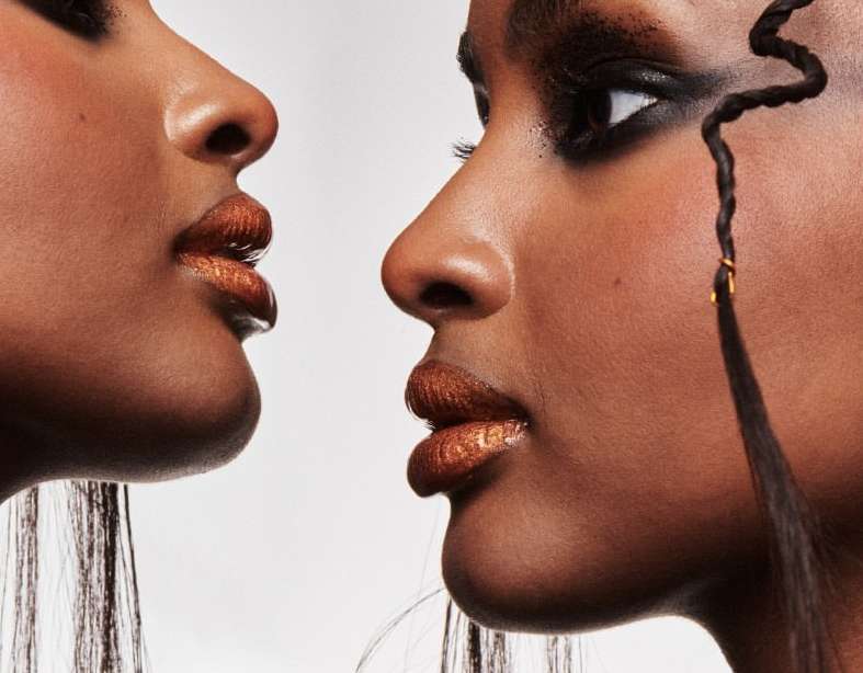 Close-up of two dark-skinned female faces, with dramatic eye makeup and shimmering lip color, set against a light neutral background.
