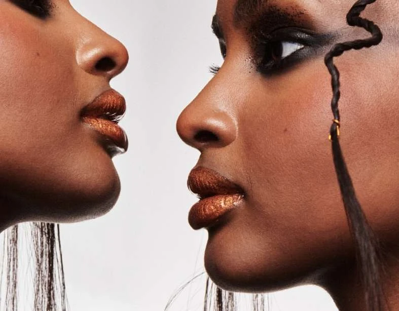 Close-up of two dark-skinned female faces, with dramatic eye makeup and shimmering lip color, set against a light neutral background.