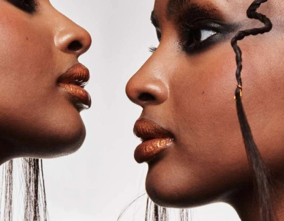 Close-up of two dark-skinned female faces, with dramatic eye makeup and shimmering lip color, set against a light neutral background.