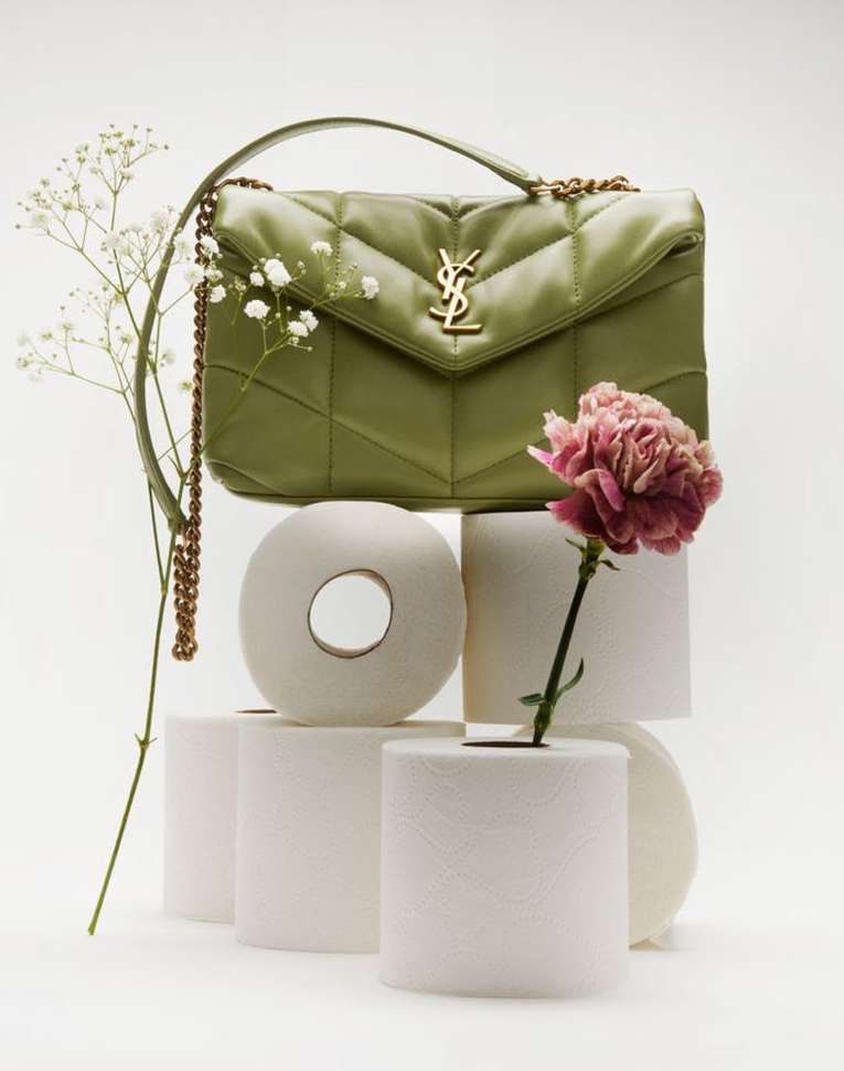 Olive-green Saint Laurent purse sits atop stacked toilet paper rolls, accented by white and pink flowers in a studio setting.
