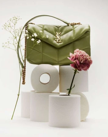 Olive-green Saint Laurent purse sits atop stacked toilet paper rolls, accented by white and pink flowers in a studio setting.