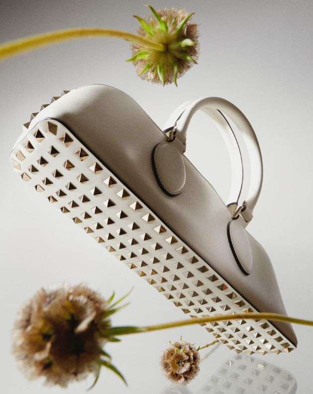 Stylish white handbag with gold stud accents, surrounded by soft, out-of-focus floral elements against a neutral background in studio lighting.